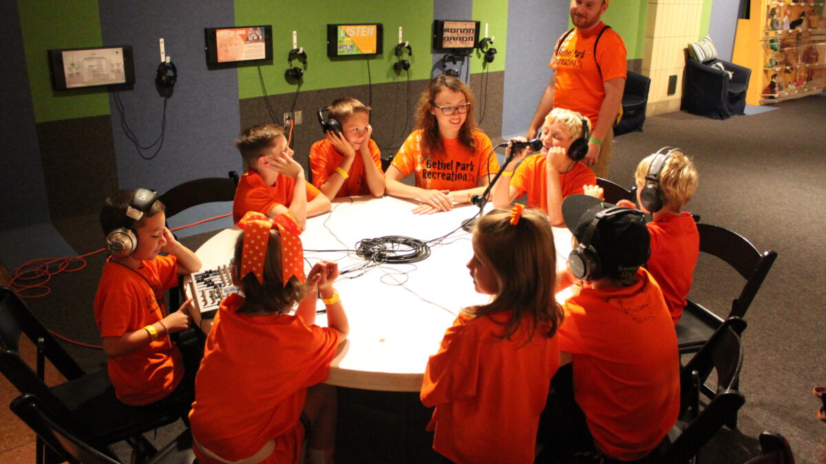 Nine youth sitting around a white table looking at an adult, five of the youth wearing headphones