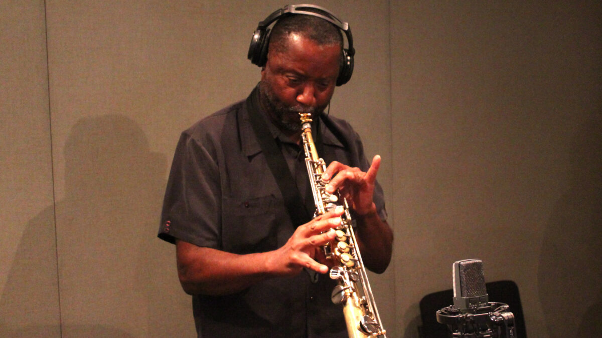 An adult wearing headphones playing a soprano saxophone into a microphone in a radio studio