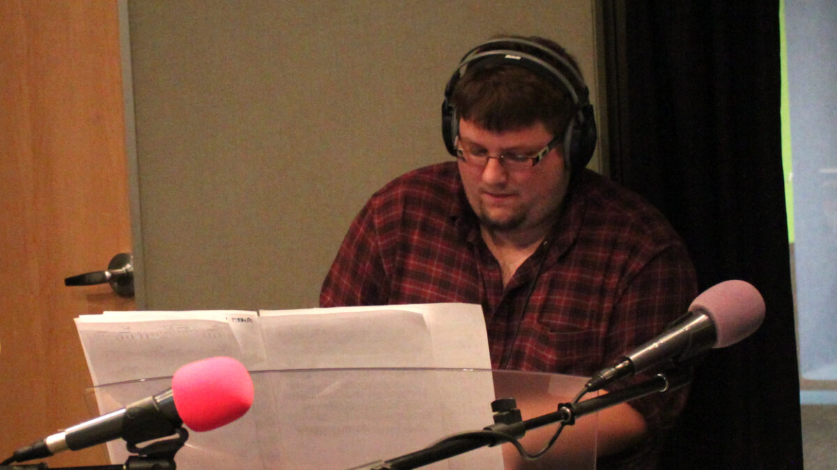 An adult wearing headphones playing a piano keyboard sitting behind a microphone in a radio studio