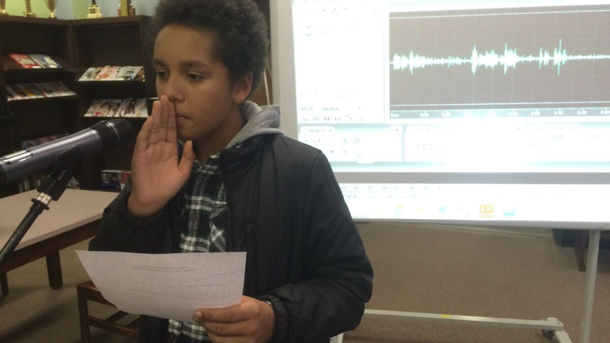 A youth holding a piece of paper standing behind a microphone in front of a projected screen of audio editing software in a classroom