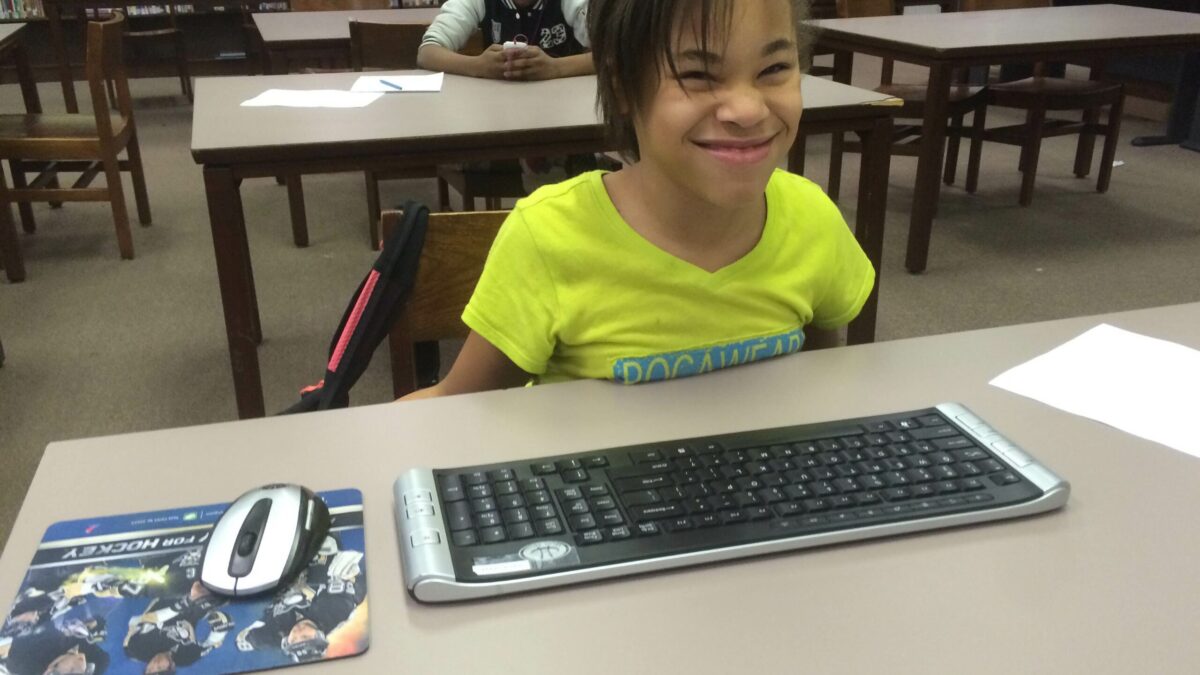 a youth smiling sitting behind a keyboard and mouse on a table, and another youth sitting in the back looking down at a phone in a library