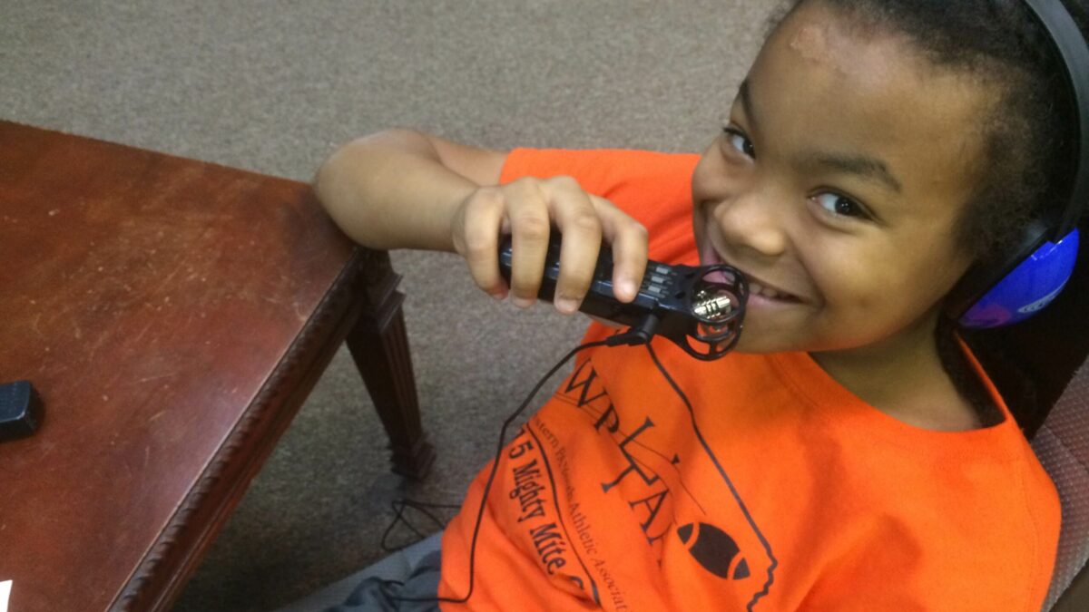 A youth wearing headphones smiling and holding a portable recorder sitting at a wooden table