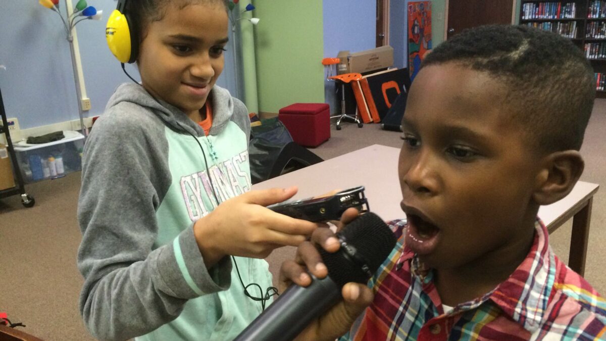 A youth wearing headphones holding a portable recorder up to another youth holding and speaking into a microphone in a classroom
