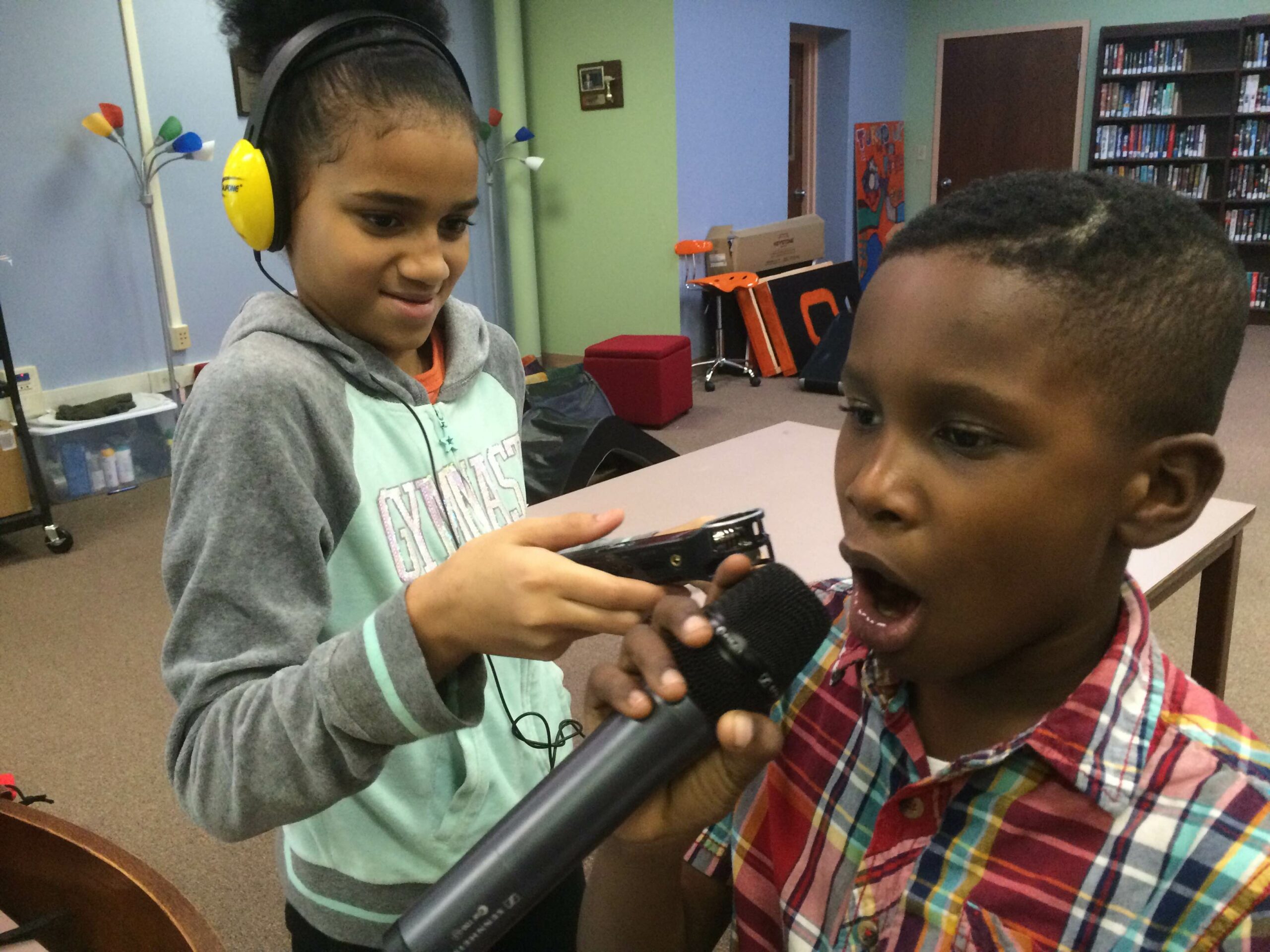 A youth wearing headphones holding a portable recorder up to another youth holding and speaking into a microphone in a classroom