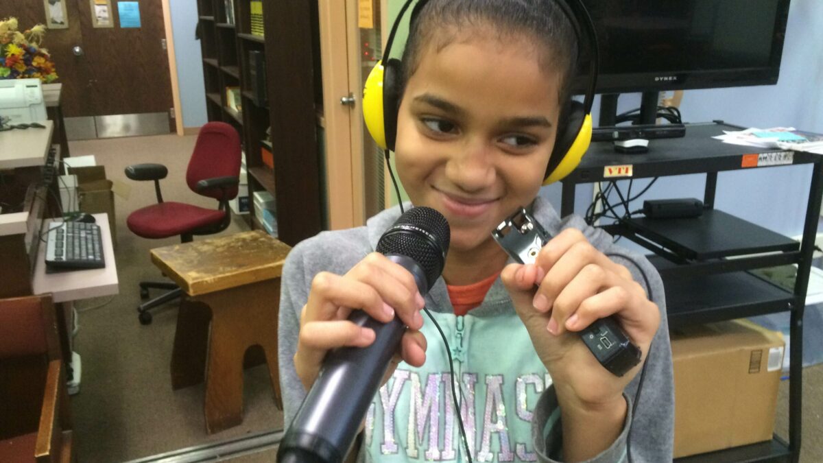 A youth wearing headphones smiling and holding a microphone and a portable recorder standing in front of a tv in a library
