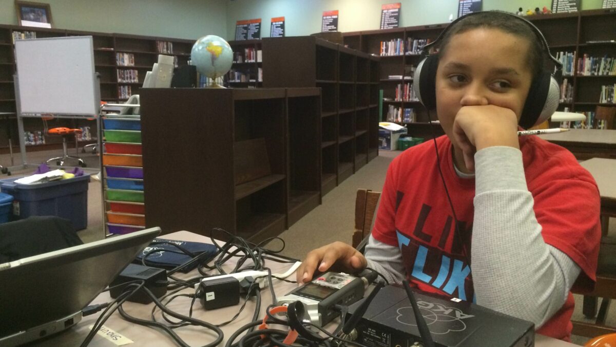 A youth wearing headphones sitting behind a portable recorder and multiple wires on a table in a library