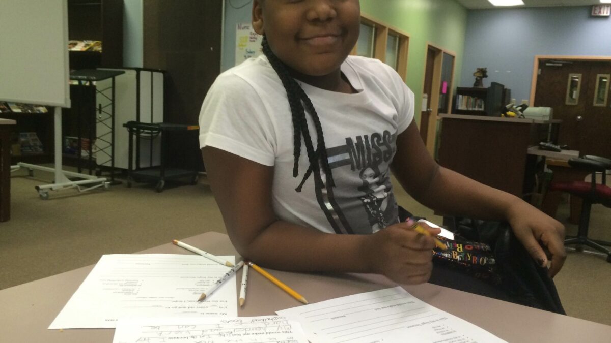 A youth smiling and sitting behind pieces of paper on a table in a library