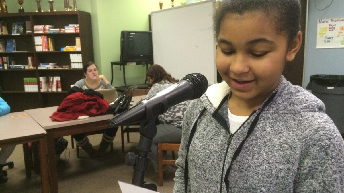 A youth holding a piece of paper speaking into a microphone as adults in the background listen in a library