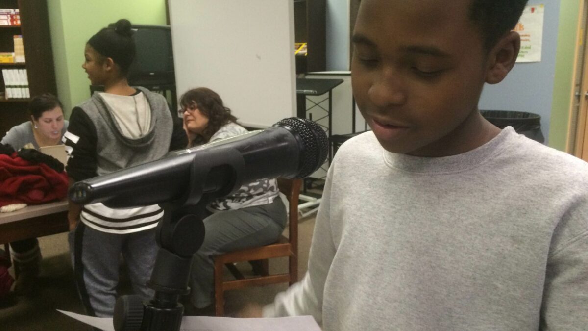 A youth reading from a piece of paper into a microphone as two adults and a youth listen in the background