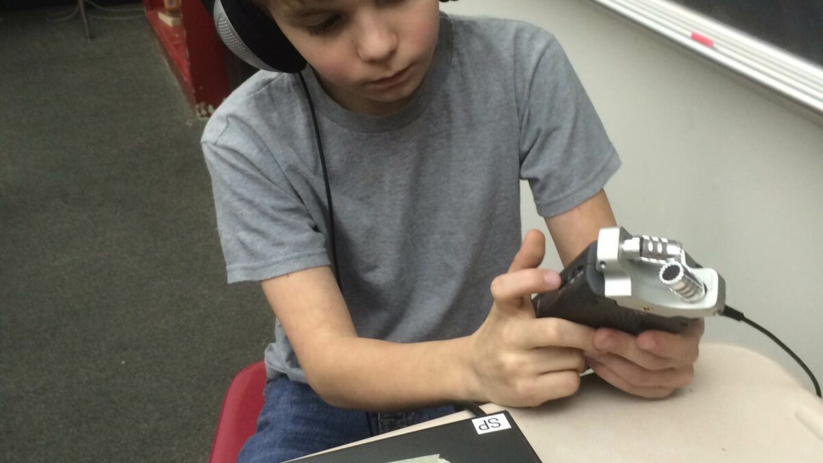 A youth wearing headphones holding a portable recorder while sitting at a desk in a classroom