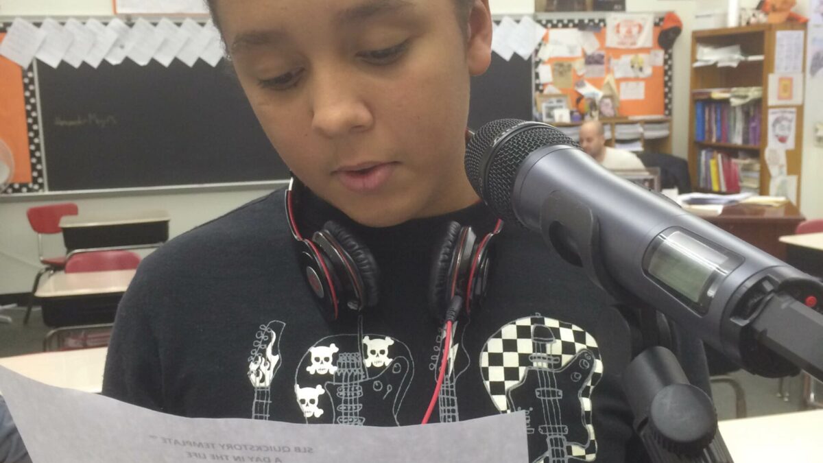 A youth reading a piece of paper and speaking into a microphone in a classroom