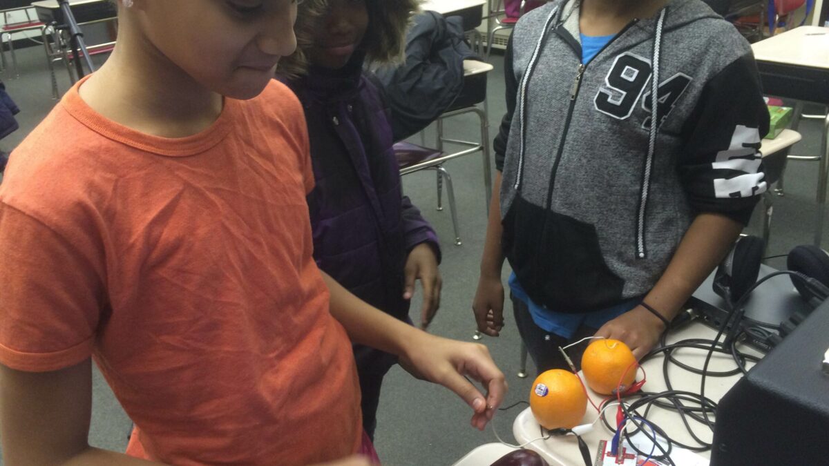 Three youth interacting with fruit connected to wires on desks in a classroom