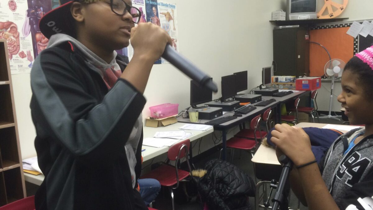 A youth singing into a microphone as another youth smiles and listens in a classroom