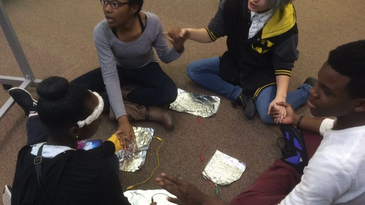 Four youth holding hands in a circle surrounding tinfoil and wires on the ground