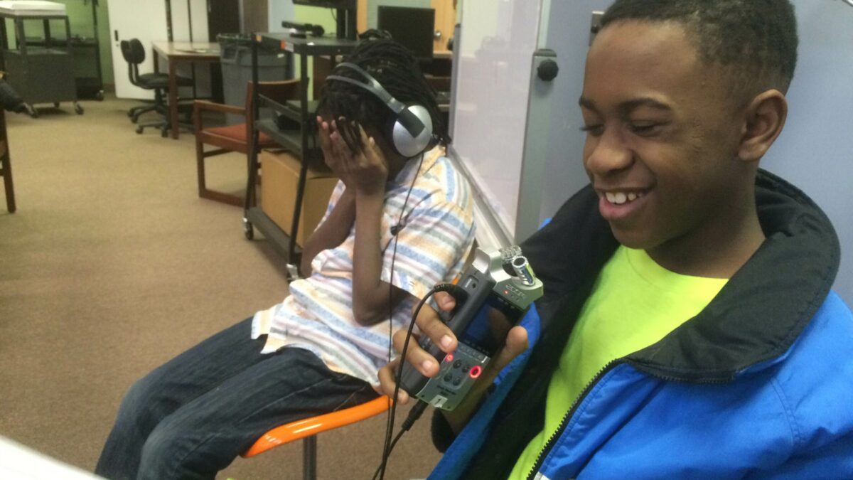 Two youth in a library, one laughing while holding a piece of paper and a recorder, the oterh wearing headphones and holding their face in their hands