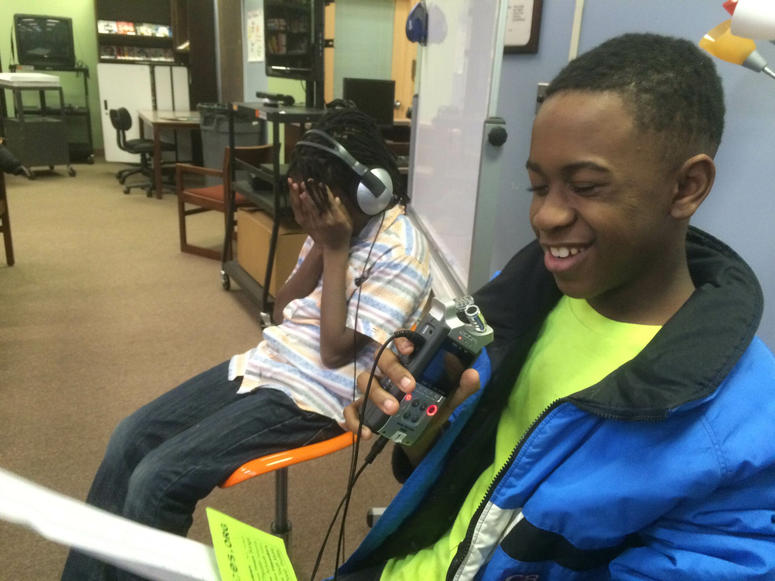 Two youth in a library, one laughing while holding a piece of paper and a recorder, the oterh wearing headphones and holding their face in their hands