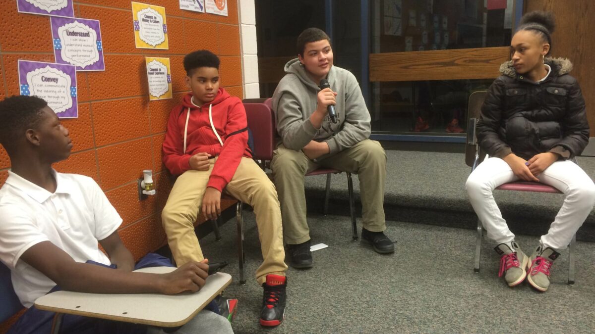 IMG_2310 Four teens sitting in chairs in a classroom, one holding and speaking into a microphone