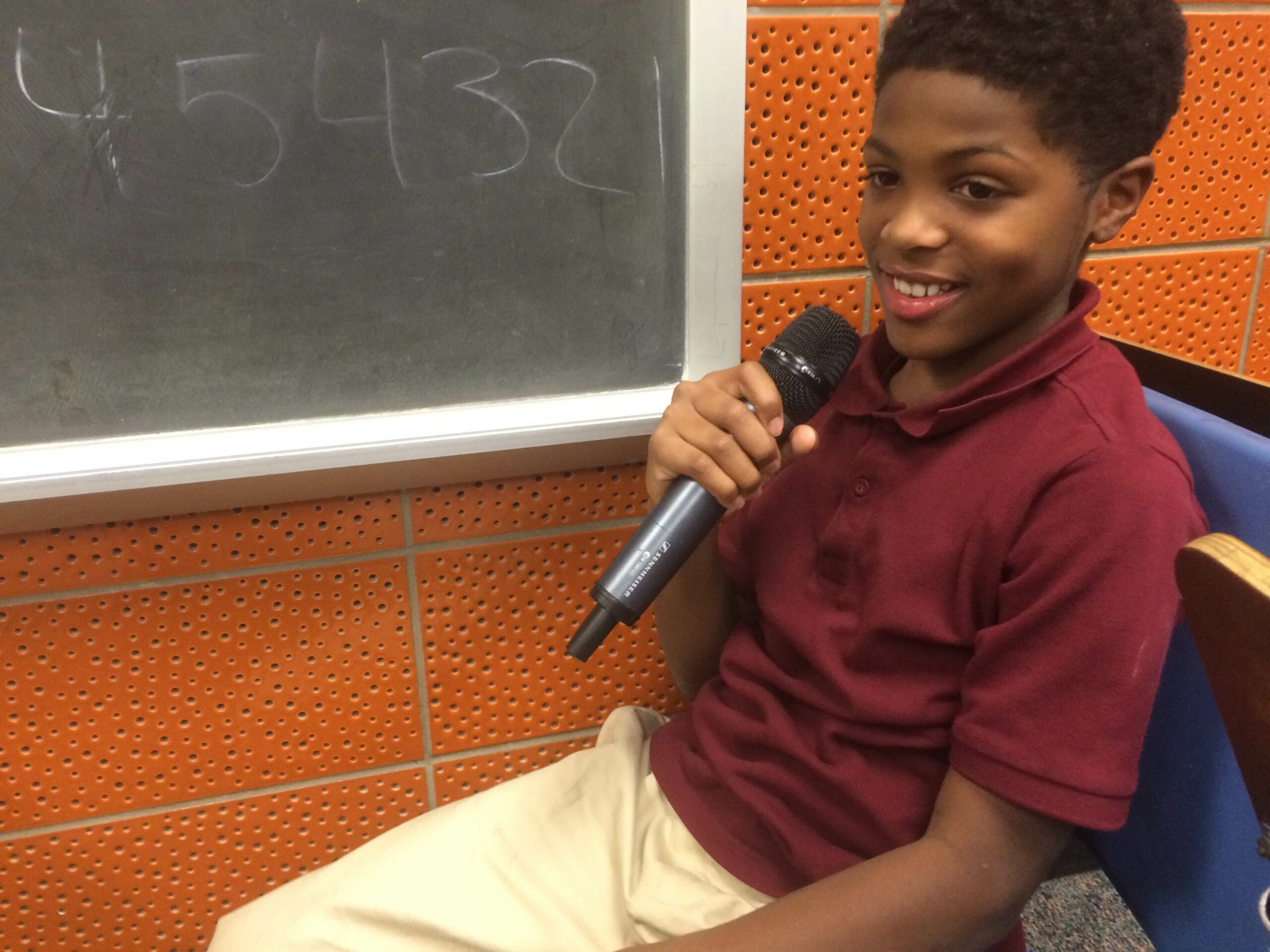 A youth holding a microphone sitting next to a blackboard in a classroom