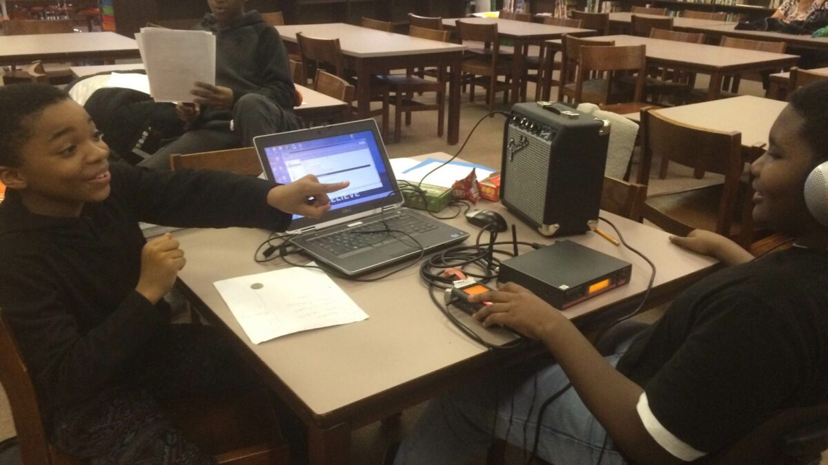 Two youth, one wearing headphones, interacting in front of a laptop computer and a portable recorder with another youth watching in the background in a library