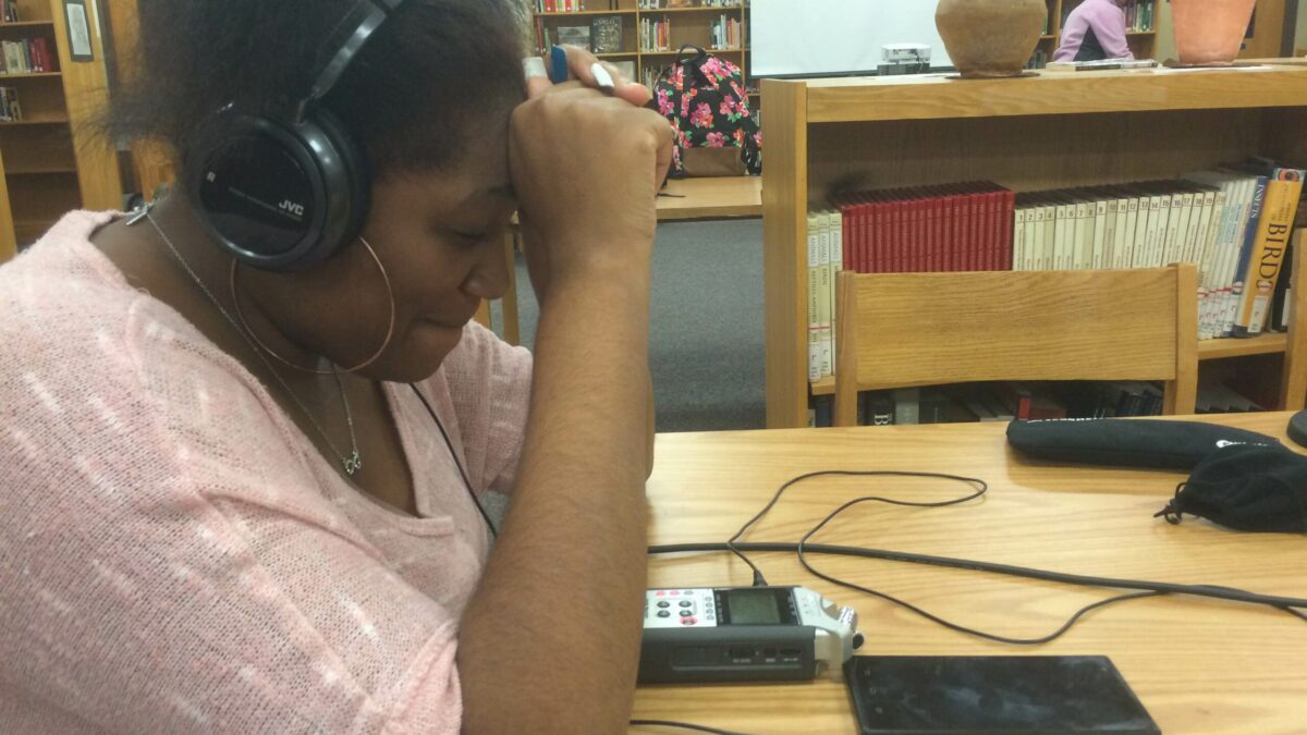 A teen wearing headphones looking down at a portable recorder and a smartphone on a wooden table in a library