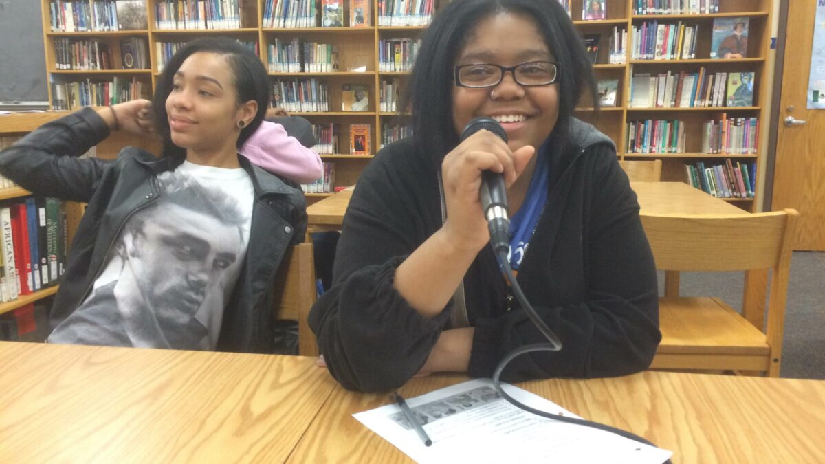 Two teens, one holding a microphone, sitting at a wooden table in a library