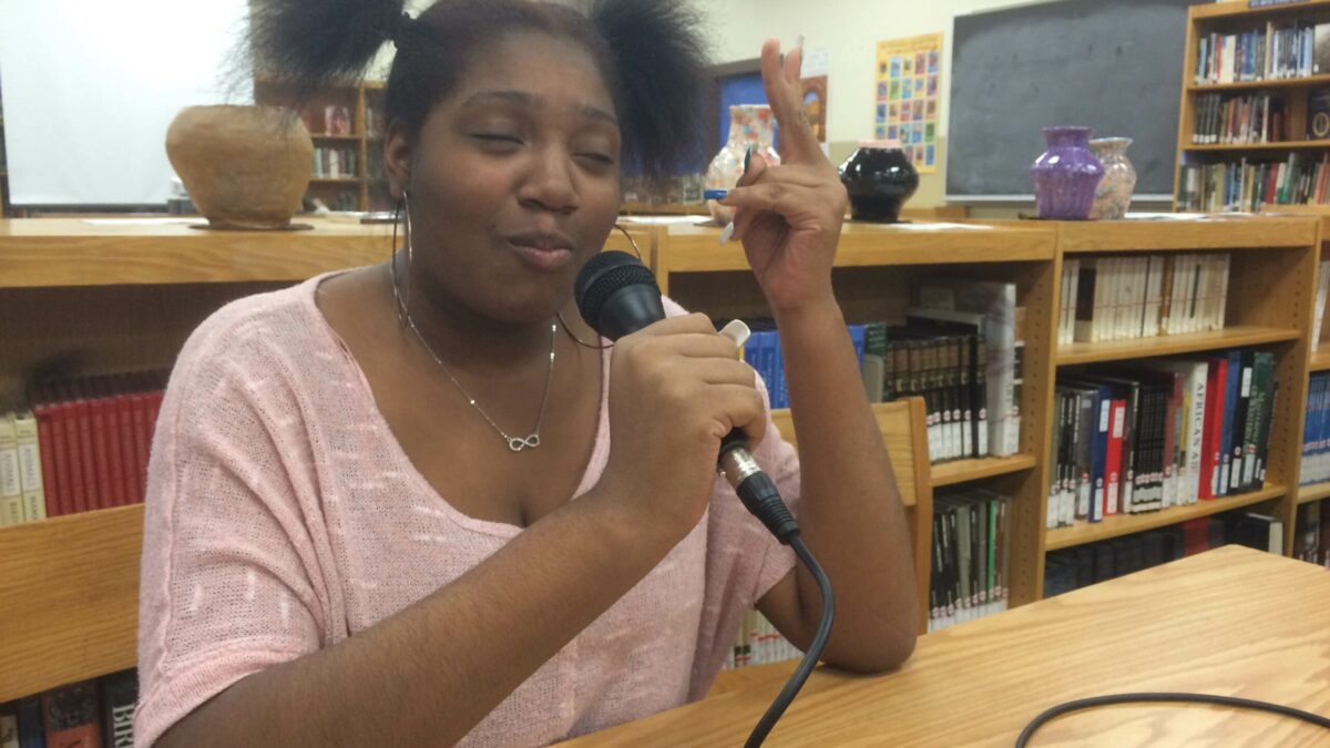 A teen holding a microphone sitting at a wooden table in a library