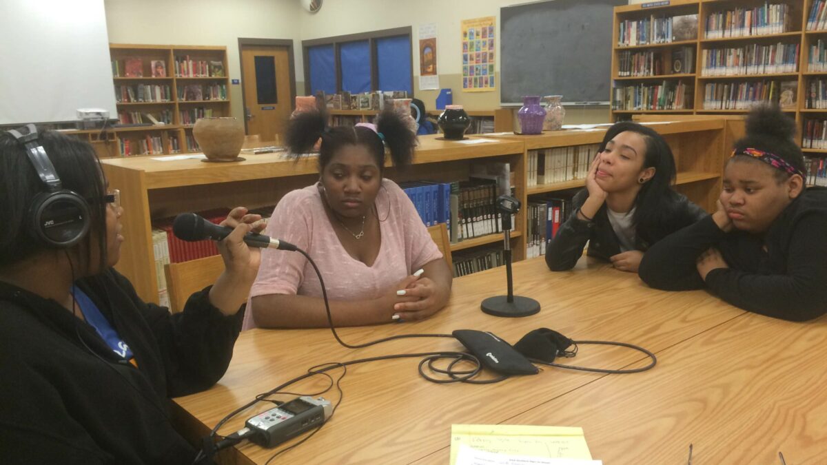 Four teens, one wearing headphones speaking into a microphone, sitting around a wooden table in a library