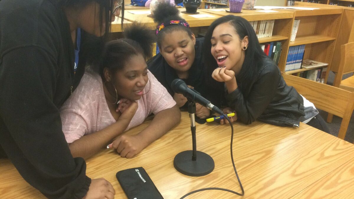 Four teens gathered around a microphone on a wooden table in a library