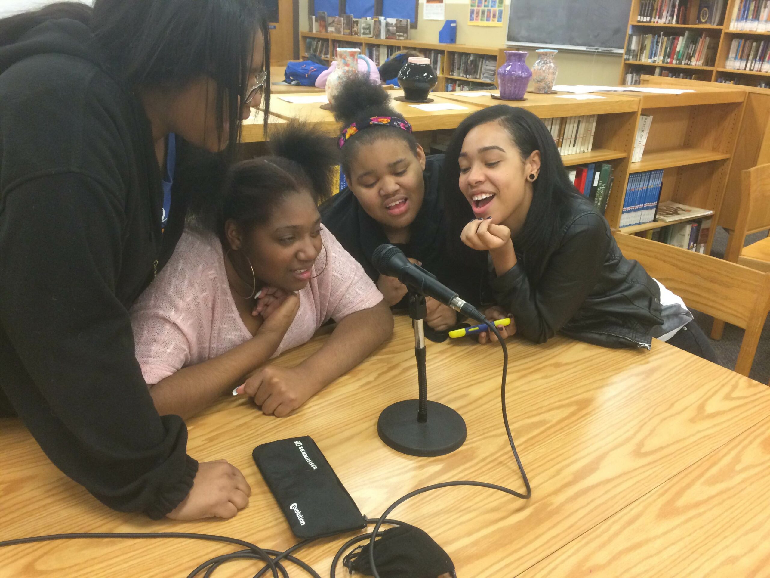 Four teens gathered around a microphone on a wooden table in a library