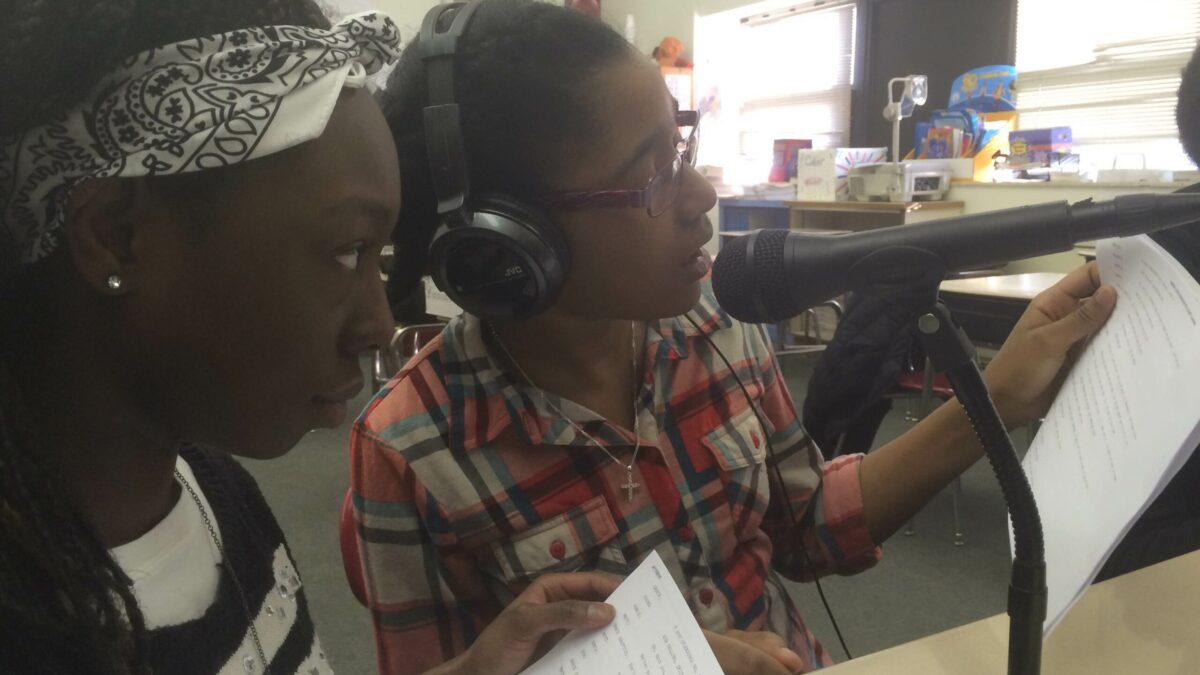 Two youth wearing headphones reading from pieces of paper into microphones