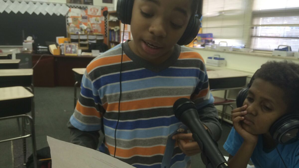 Two youth wearing headphones reading from a piece of paper into a microphone