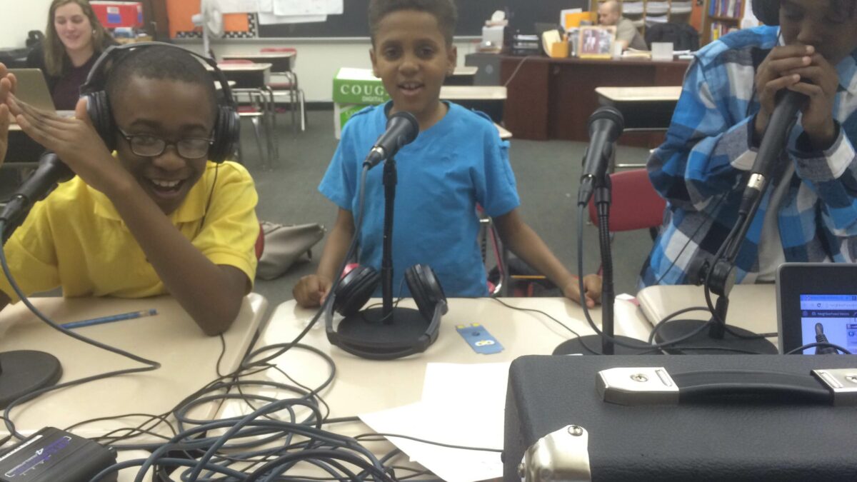 Three youth, two wearing headphones, speaking into headphones as an adult oversees in a classroom