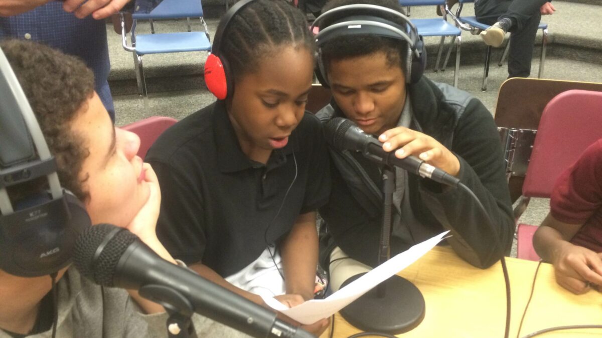 Three youth wearing headphones, one looking at a piece of paper and speaking into a microphone, sitting in a classroom