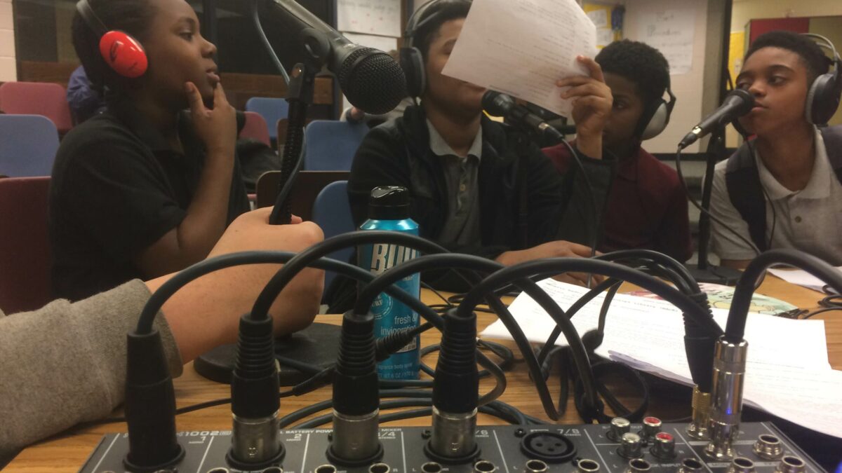 A closeup of microphones plugged into a mixer, with four youth wearing headphones sitting behind the microphones in a classroom