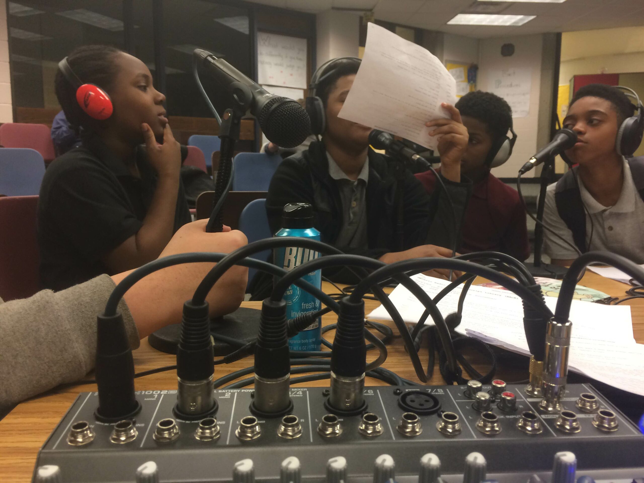 A closeup of microphones plugged into a mixer, with four youth wearing headphones sitting behind the microphones in a classroom