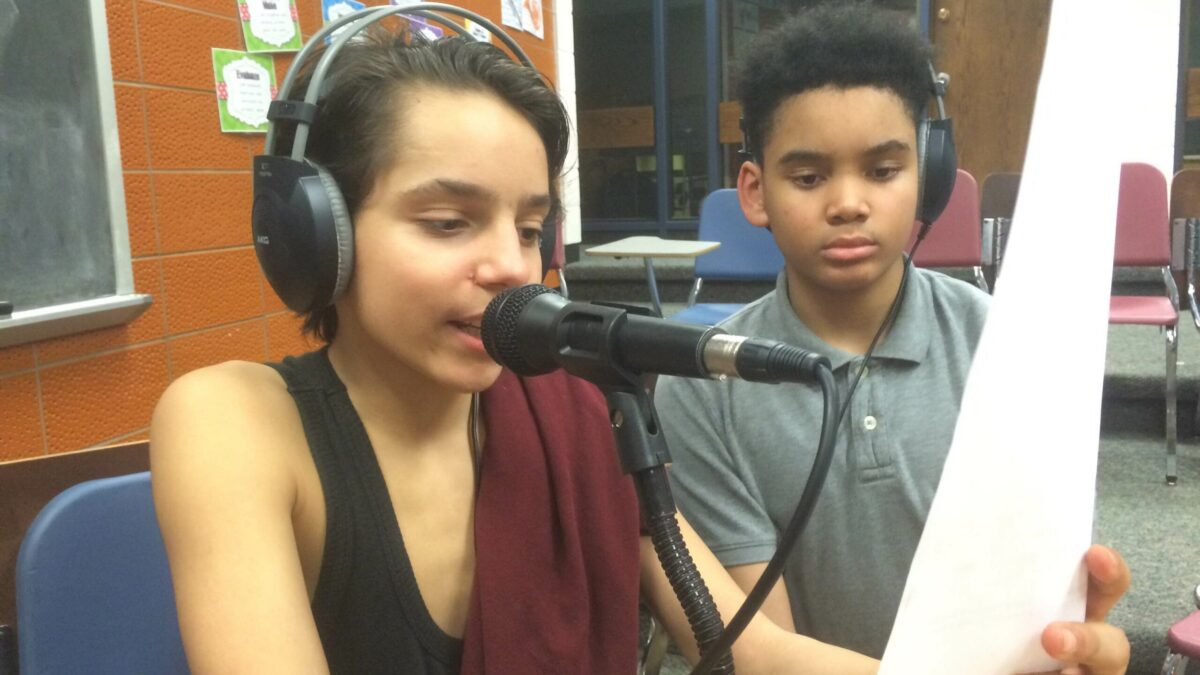 Two youth wearing headphones, one speaking into a microphone and holding up a piece of paper in a classroom