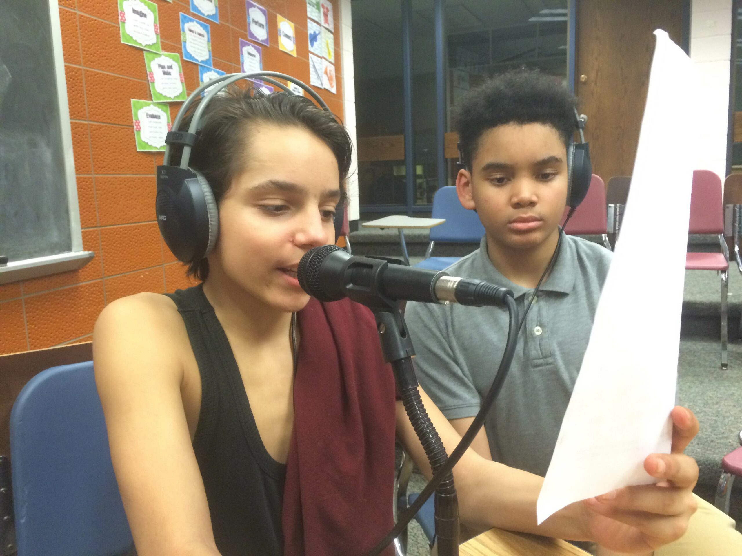 Two youth wearing headphones, one speaking into a microphone and holding up a piece of paper in a classroom