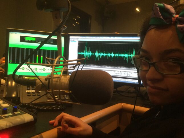 A youth looking at the camera sitting behind a microphone and two computer screens displaying audio information in a radio studio