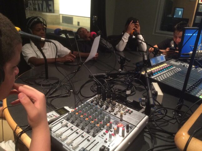 Four teens, two wearing headphones on their head, two wearing headphones around their neck, sitting behind microphones as another youth wearing headphones looks down at a mixer in a radio studio