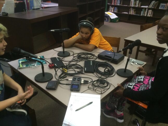 Three youth wearing headphones speaking into microphones sitting around a square table in a library