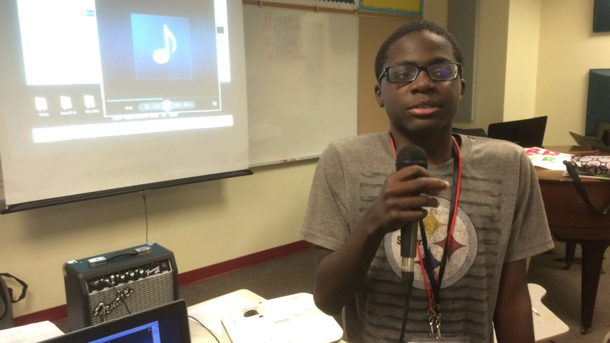 A youth holding a microphone standing next to a laptop computer and in front of a projector in a classroom