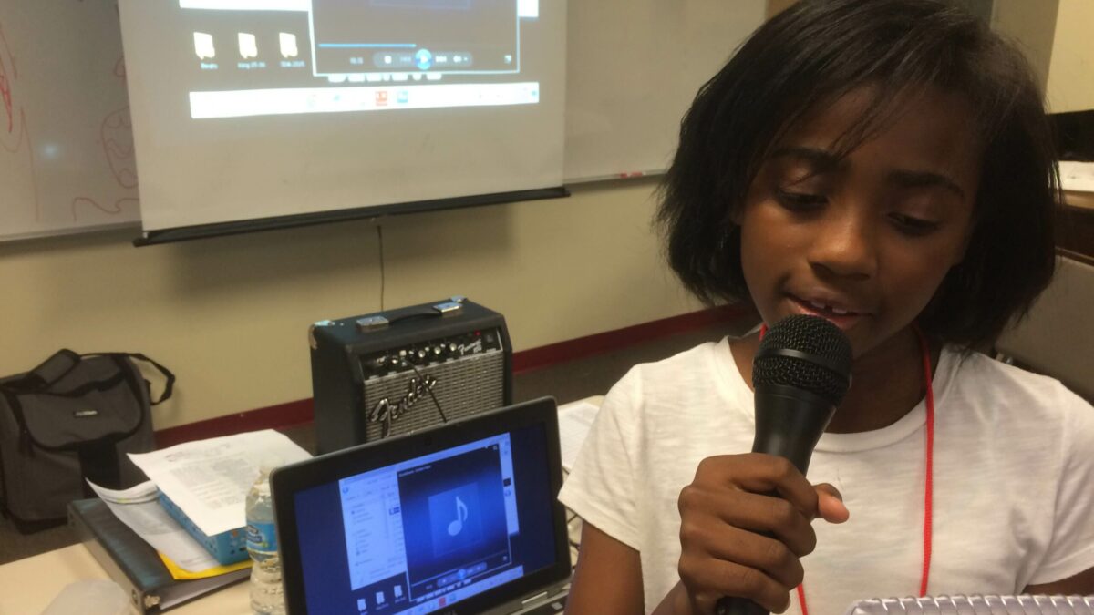 A youth holding a notepad speaking into a microphone standing in front of a laptop computer in a classroom