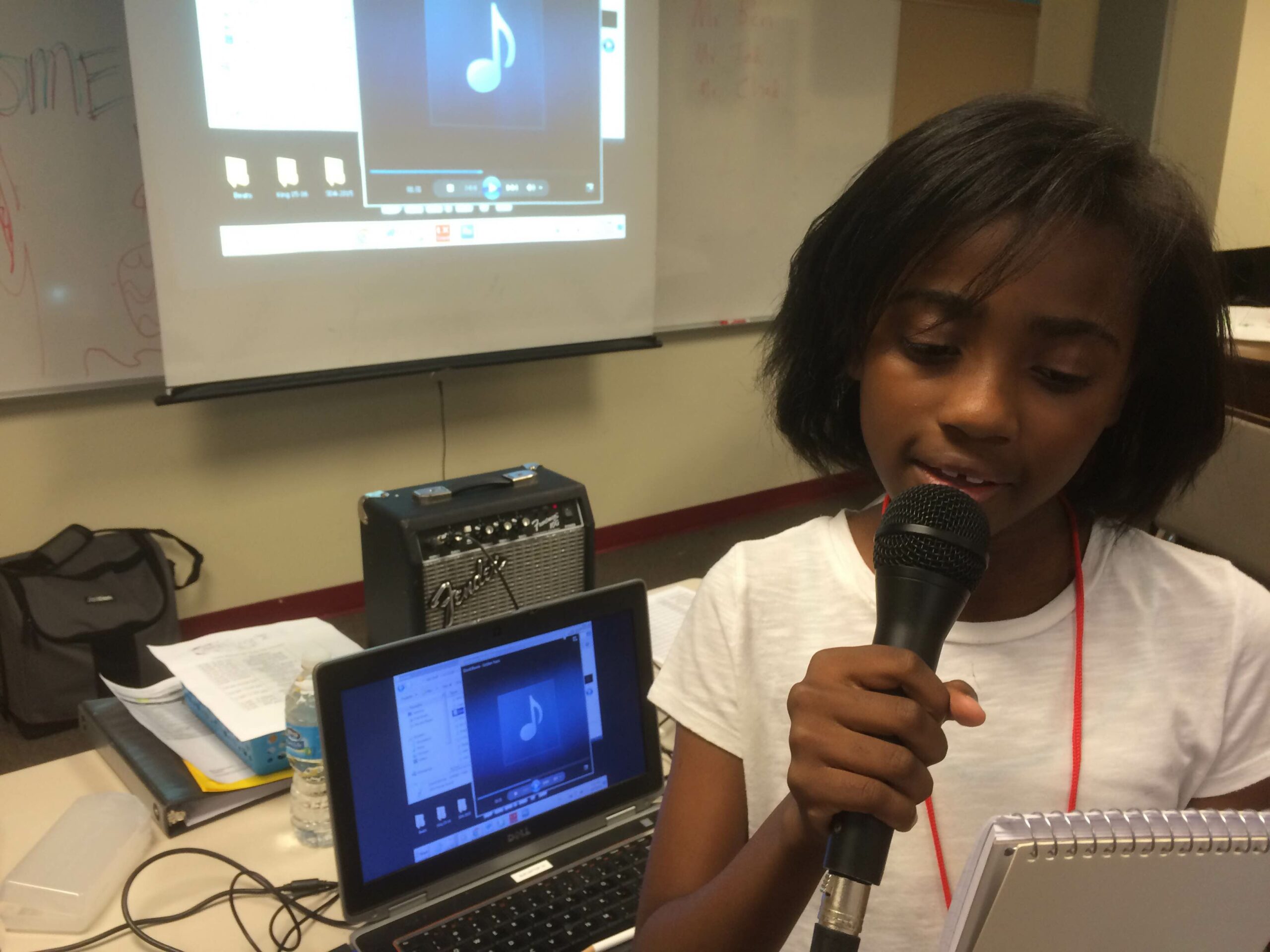 A youth holding a notepad speaking into a microphone standing in front of a laptop computer in a classroom