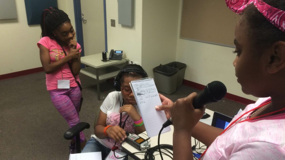 A youth holding a notepad and a microphone, a youth wearing headphones looking down at a portable recorder, and another youth standing nearby