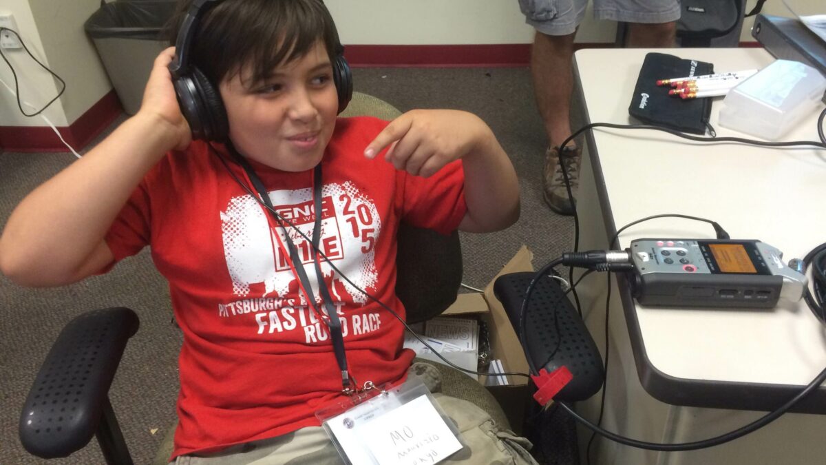 A youth wearing headphones sitting at a desk with a portable recorder on it in a classroom