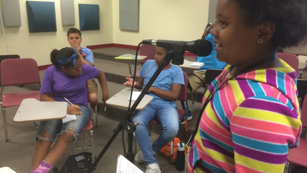 IMG_2970 A youth holding a notepad and speaking into a microphone and three youth sitting at classroom desks listening