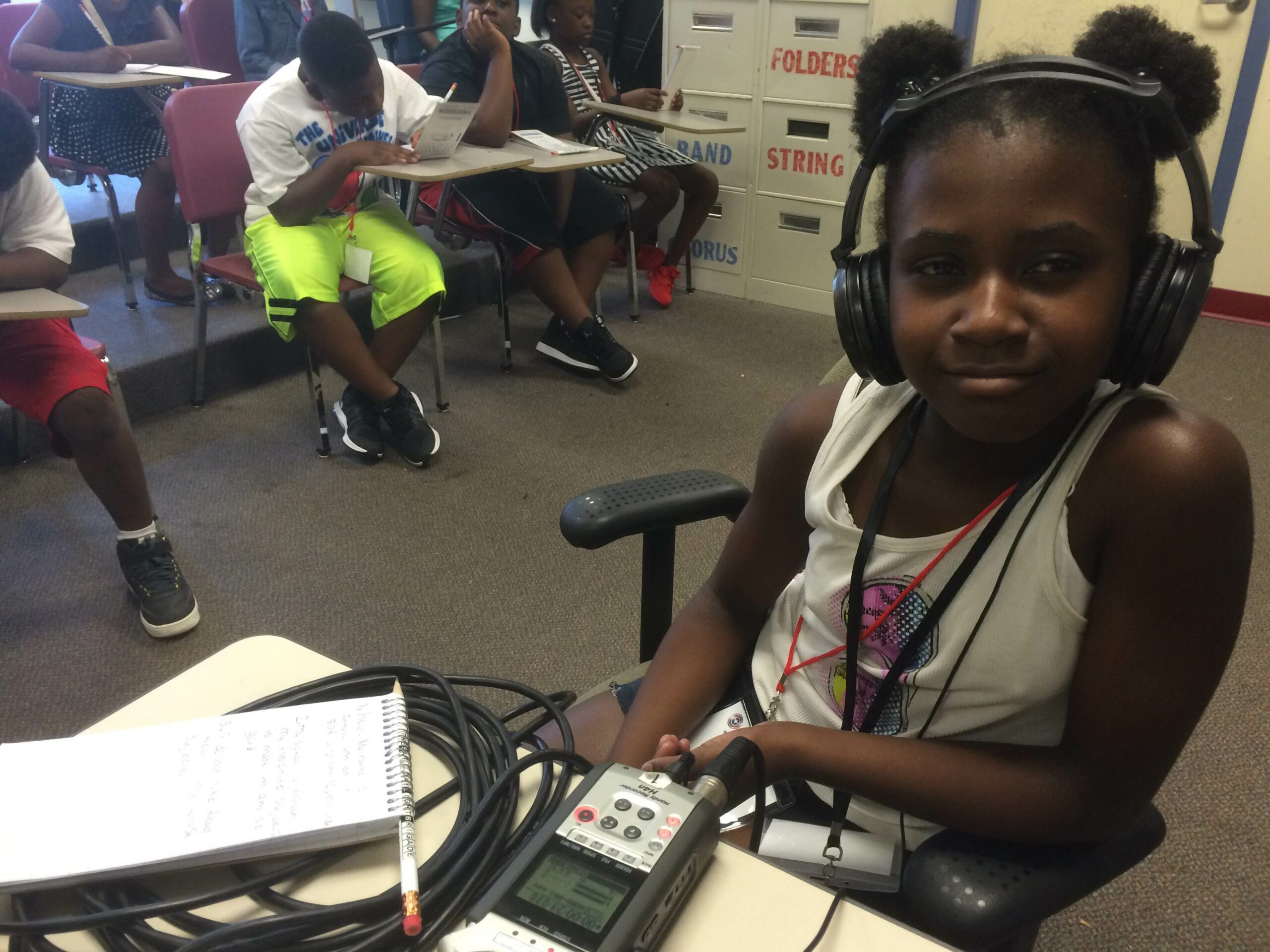 A youth wearing headphones sitting behind a portable microphone and a notepad on a desk in a classroom