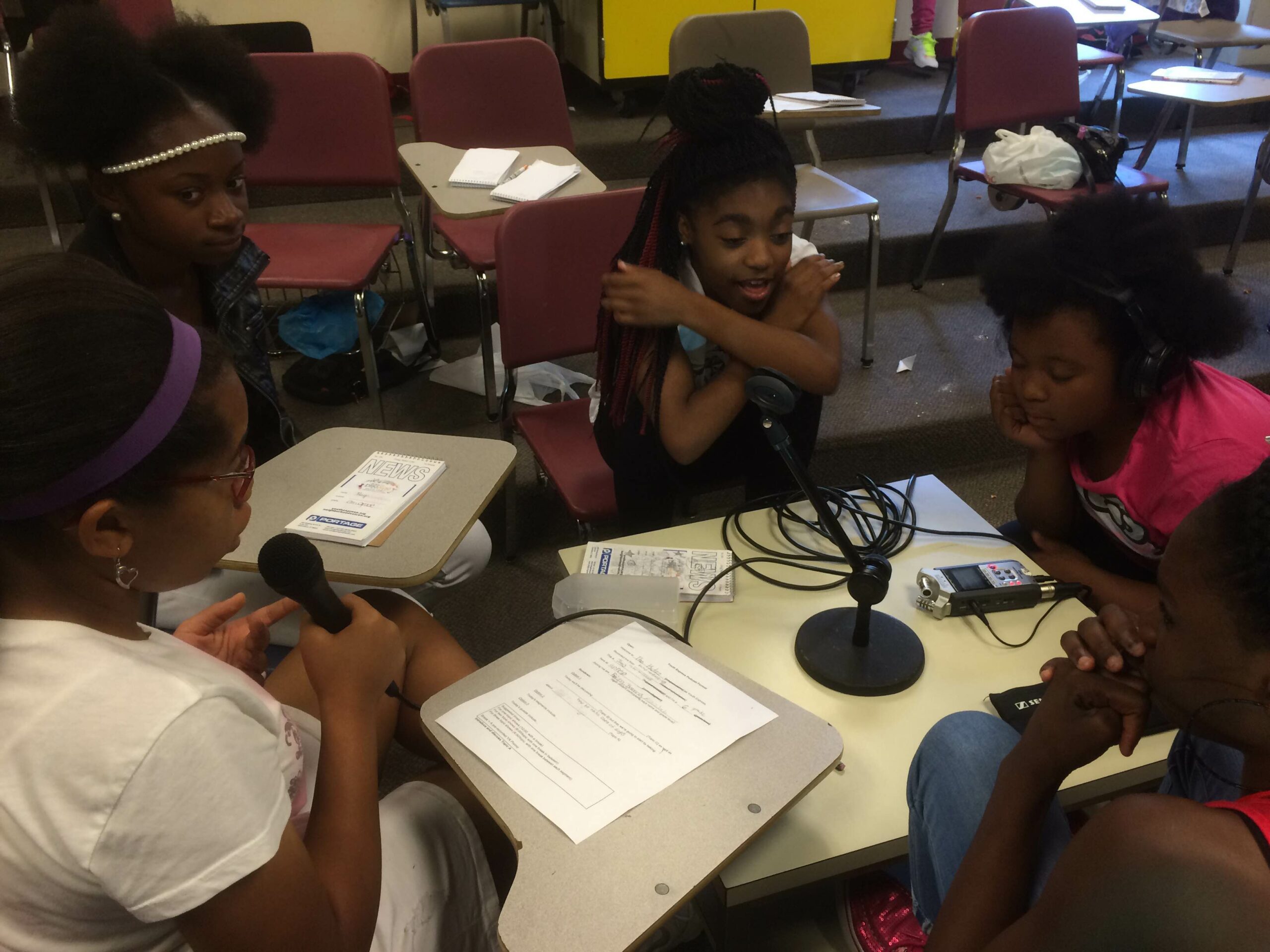 Three youth sitting around a portable recorder on a table, one youth holding a microphone