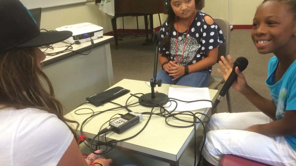 Three youth sitting around a portable recorder, one wearing headphones, one holding a microphone and smiling