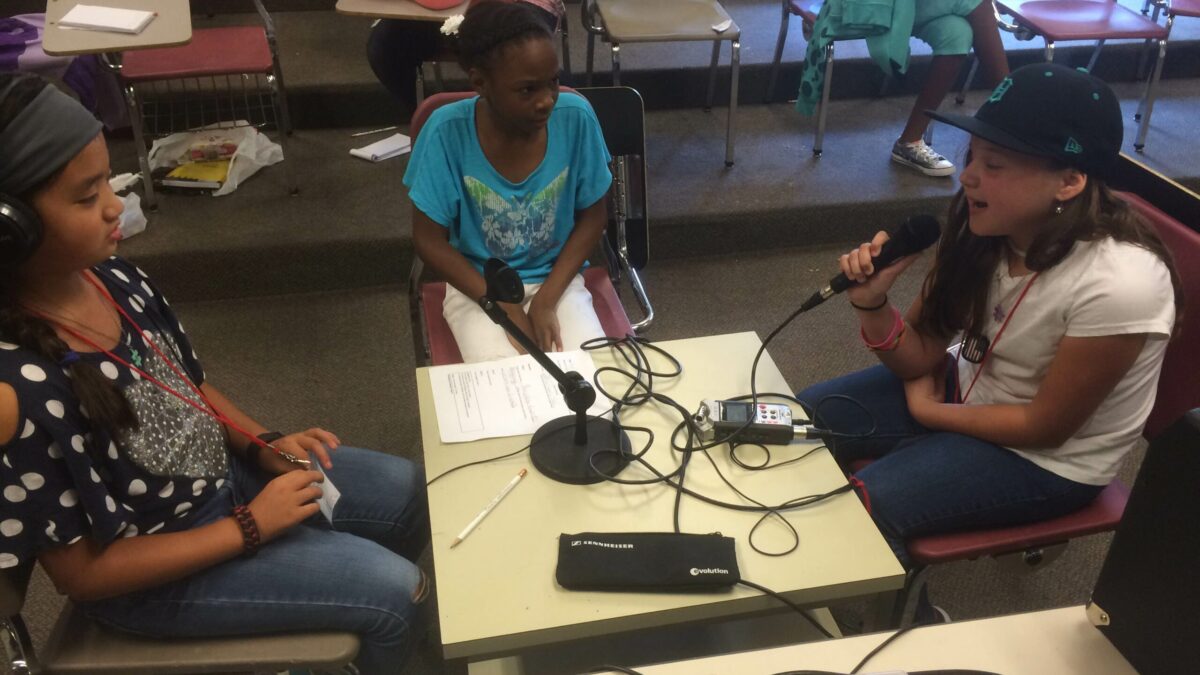 Three youth sitting around a portable recorder, one wearing headphones, one holding a microphone and smiling
